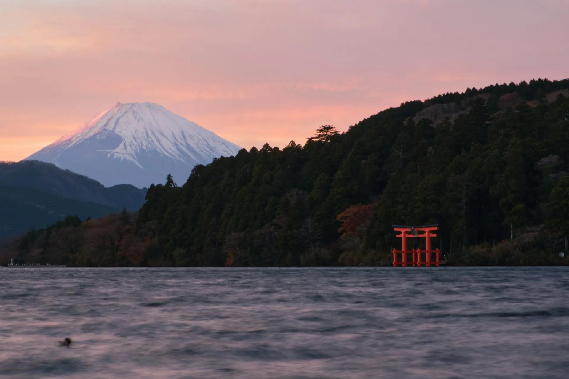 Monte Fuji al tramonto visto dal lago Ashi, con torii rosso sull’acqua, boschi sulla riva e cielo rosato sullo sfondo.