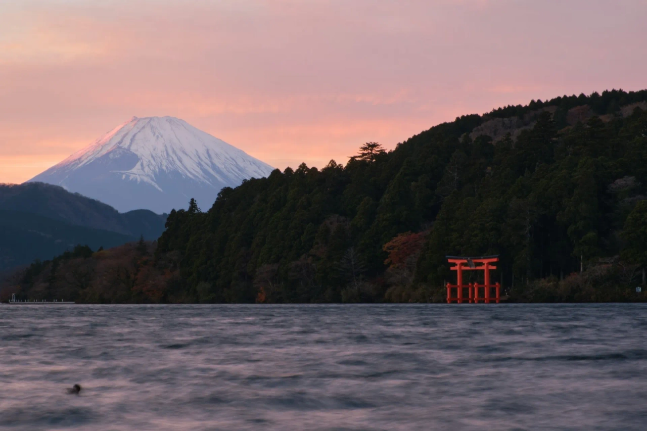 Monte Fuji al tramonto visto dal lago Ashi, con torii rosso sull’acqua, boschi sulla riva e cielo rosato sullo sfondo.