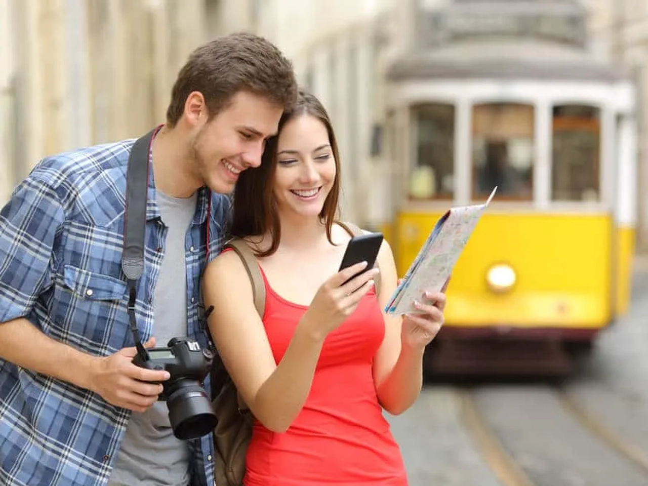 Casal sorridente usando smartphone e mapa em uma rua com bonde amarelo.