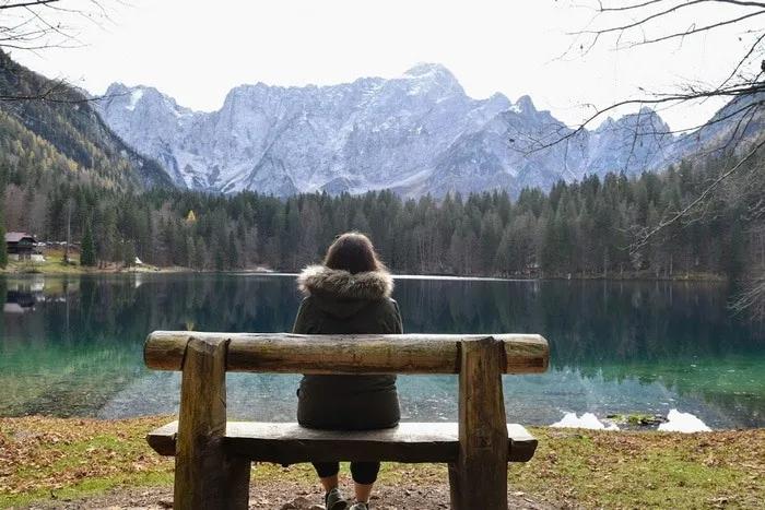 Persona sentada en un banco, contemplando un lago rodeado de montañas y árboles.