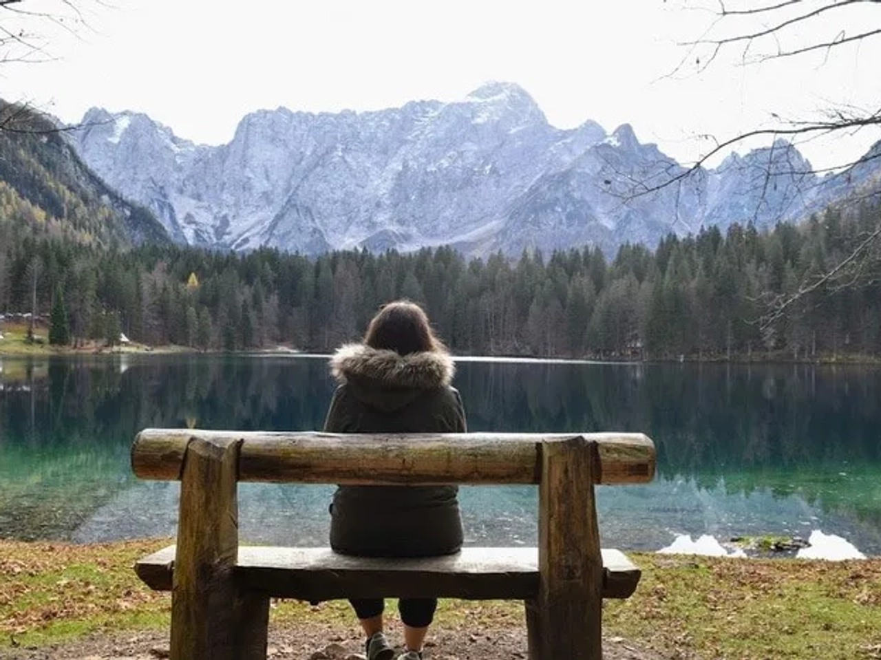 Persona sentada en un banco, contemplando un lago rodeado de montañas y árboles.