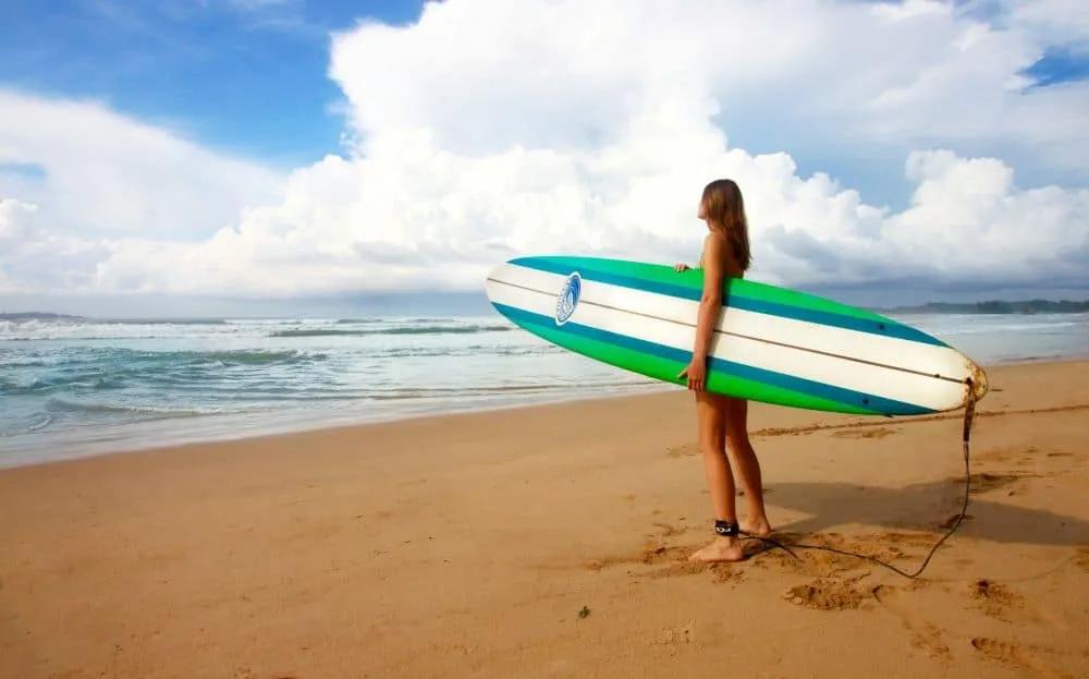 Joven con tabla de surf en la playa, mirando el océano y el cielo.