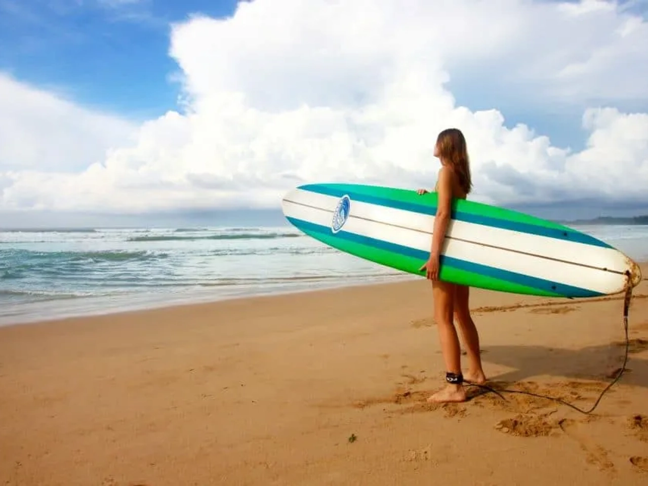 Joven con tabla de surf en la playa, mirando el océano y el cielo.