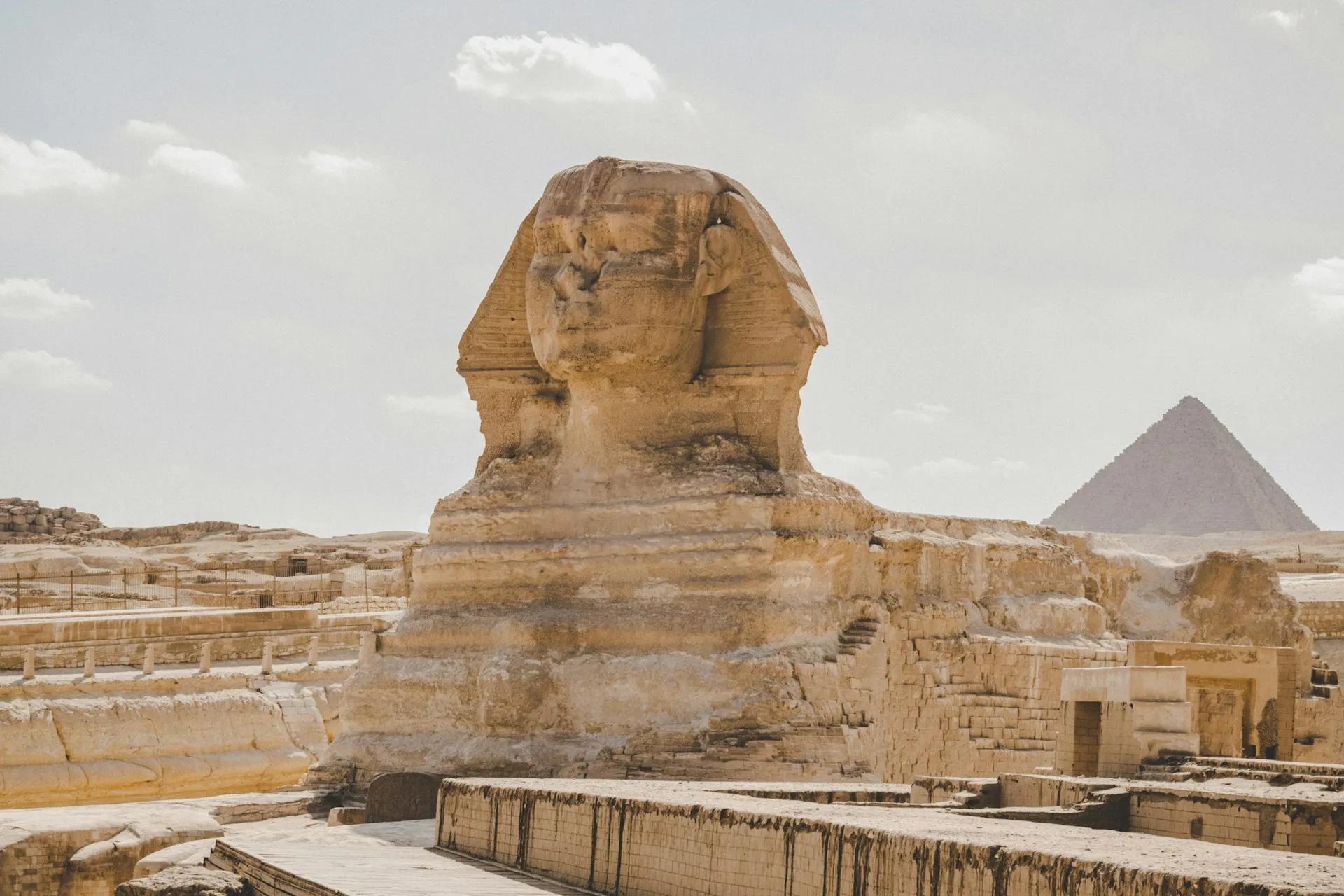 Grande Sfinge di Giza vista di lato nel complesso archeologico egiziano, con una piramide sullo sfondo a destra e rovine in pietra in primo piano.
