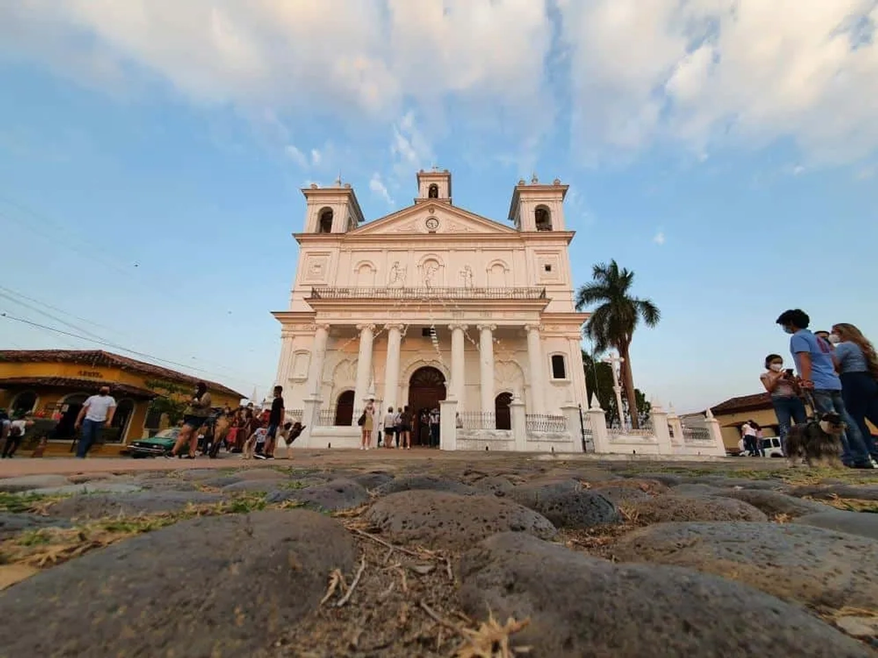 Iglesia blanca con columnas, rodeada de personas y un cielo azul al atardecer.