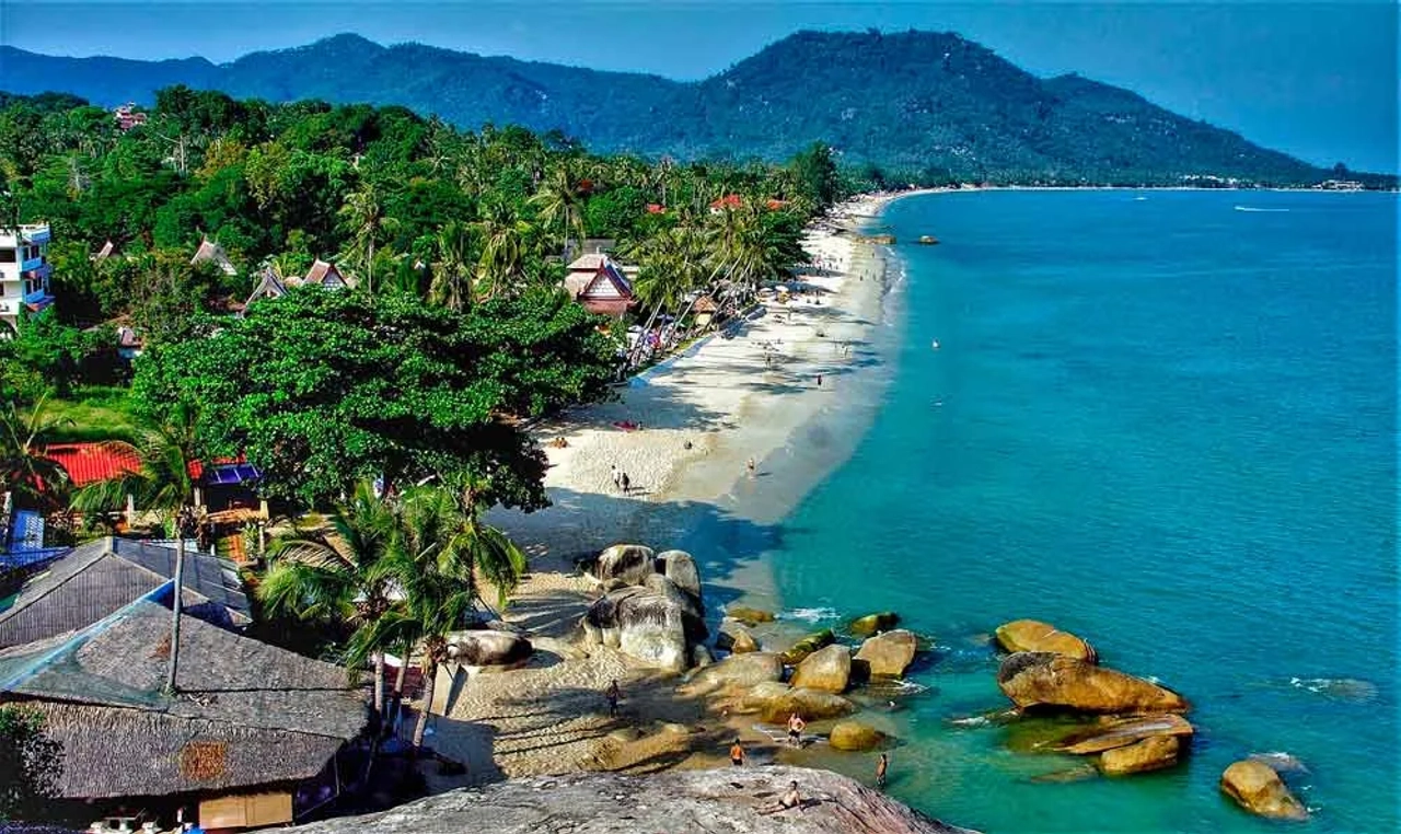 Vista panoramica di una spiaggia a Koh Samui, con mare turchese, palme, rocce sulla riva e montagne verdi sullo sfondo.