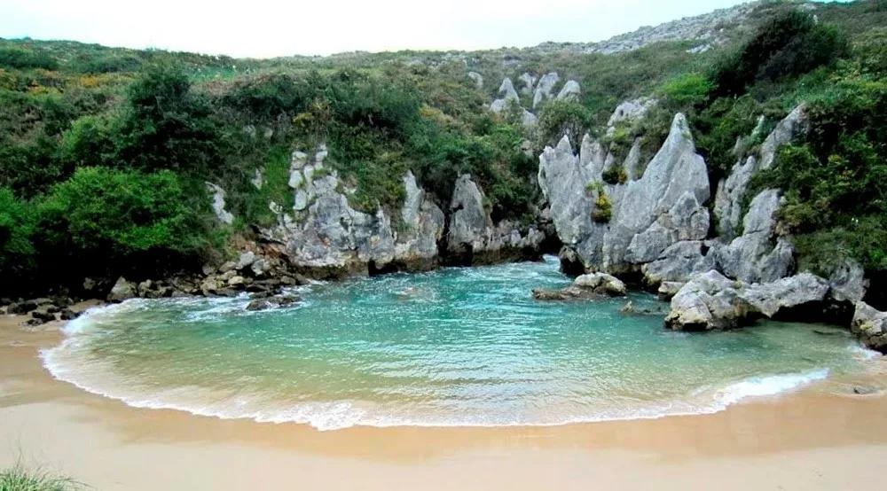 Praia tranquila cercada por rochas e vegetação nas Astúrias. Água cristalina.