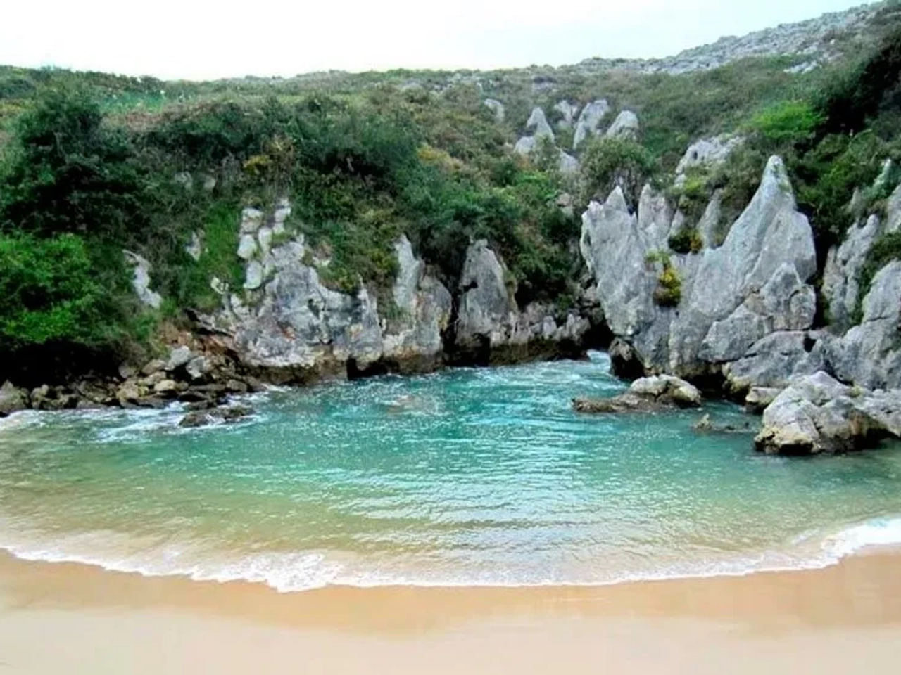 Praia tranquila cercada por rochas e vegetação nas Astúrias. Água cristalina.