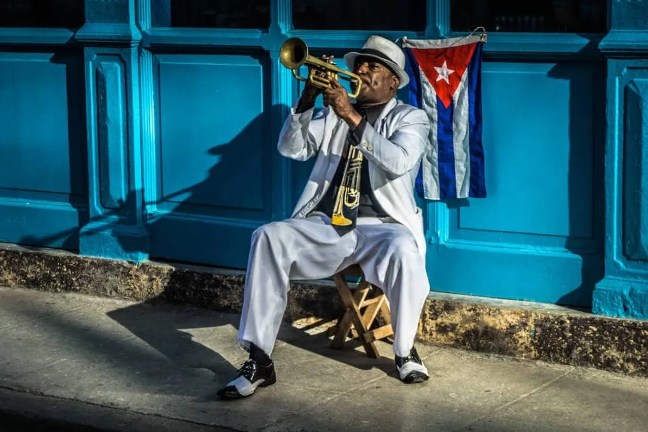 homem de fato a tocar trompeta com bandeira cubana por trás