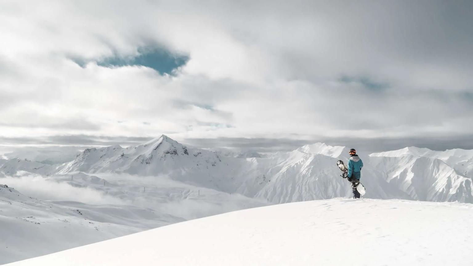 A snowboarder stands on a snowy peak, overlooking distant mountains under a cloudy sky.