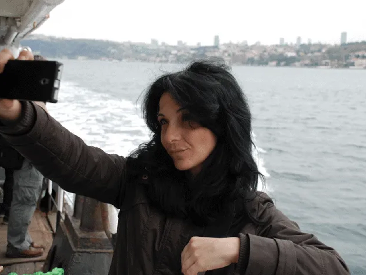 Mujer tomando una selfie en un barco con paisaje urbano al fondo.