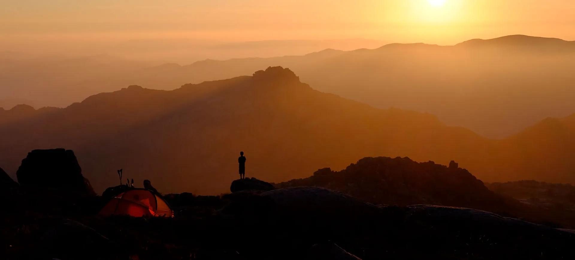 Pôr do sol sobre montanhas com uma pessoa ao lado de uma tenda.