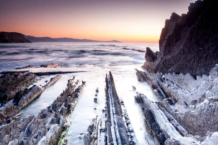 Rocas costeras al atardecer con mar tranquilo y horizonte montañoso.