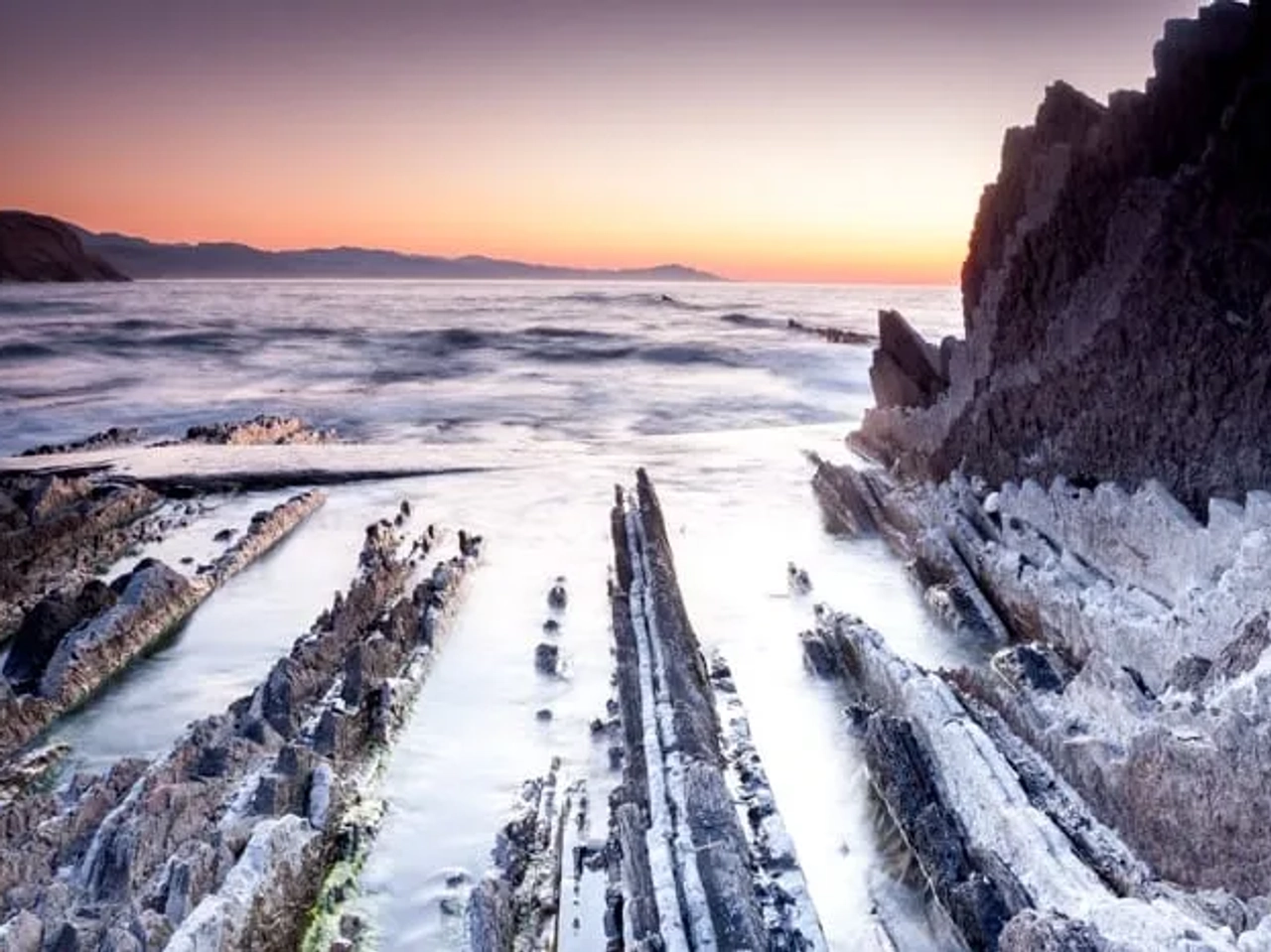 Rocas costeras al atardecer con mar tranquilo y horizonte montañoso.
