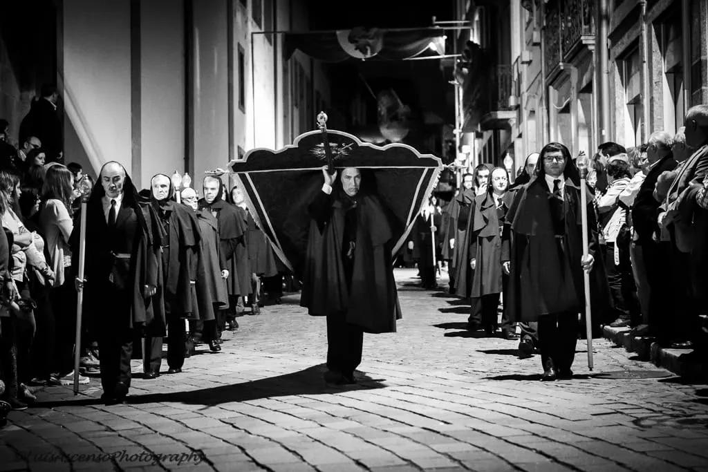 Procissão da Semana Santa com participantes vestidos de preto em uma rua histórica.