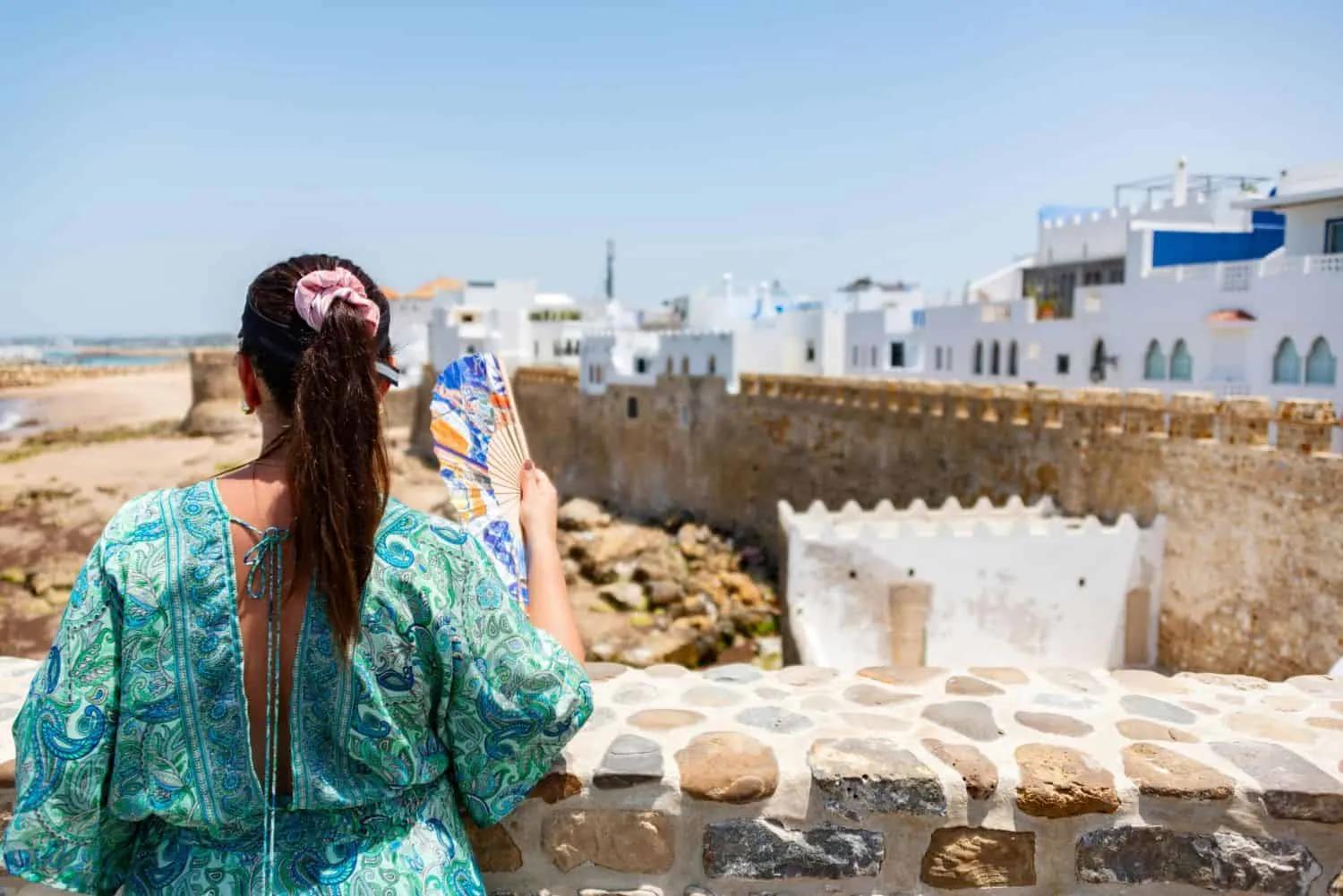 Mulher admirando a vista de uma praia em Marrocos, com casas brancas ao fundo.