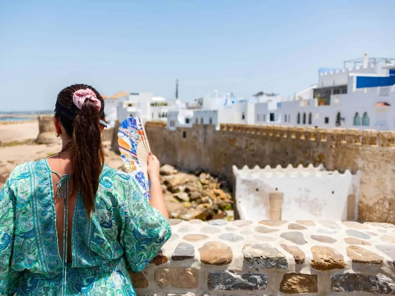 Mulher admirando a vista de uma praia em Marrocos, com casas brancas ao fundo.