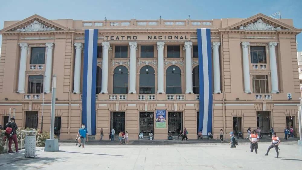 Teatro Nacional de El Salvador, edificio histórico con banderas y personas en la plaza.