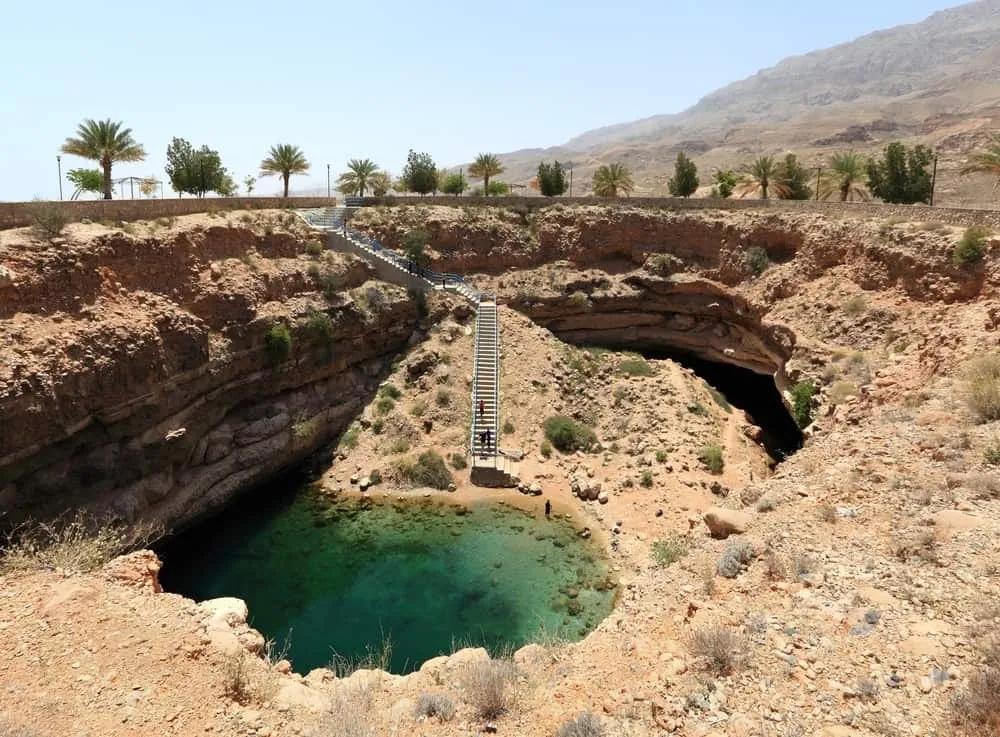 Escalera que desciende a un pozo natural rodeado de montañas y palmeras.