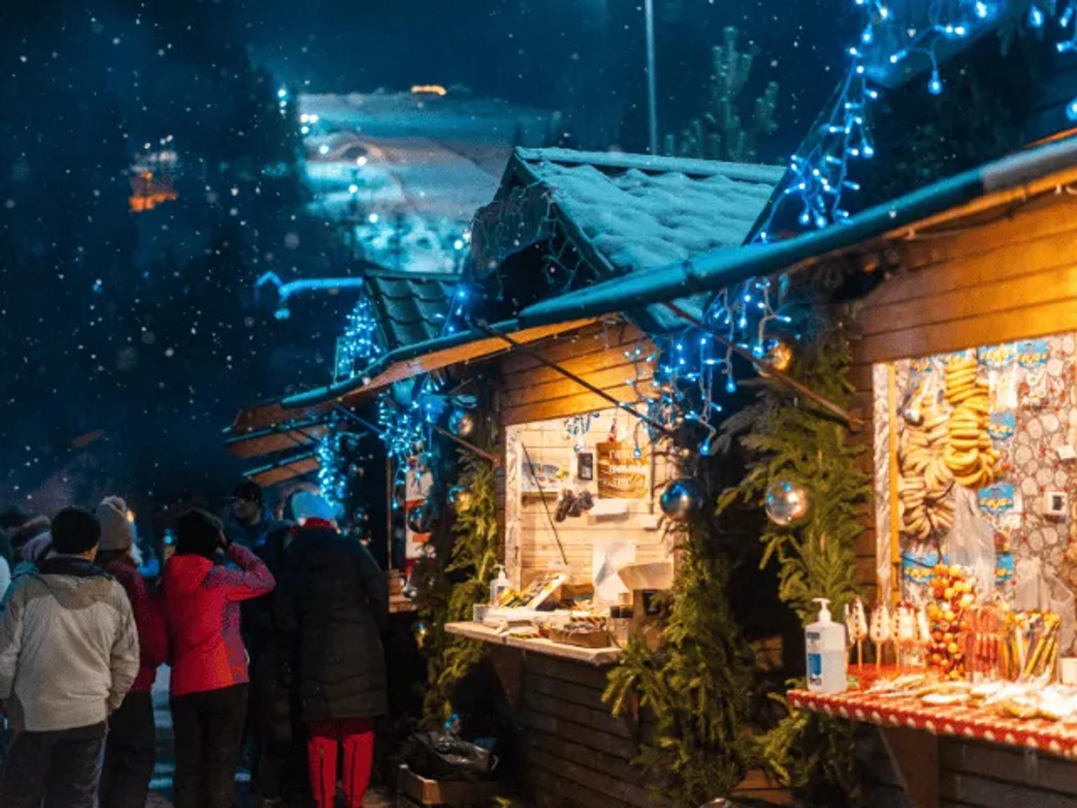 Mercado de Natal iluminado, com barracas decoradas e visitantes em um ambiente nevado.
