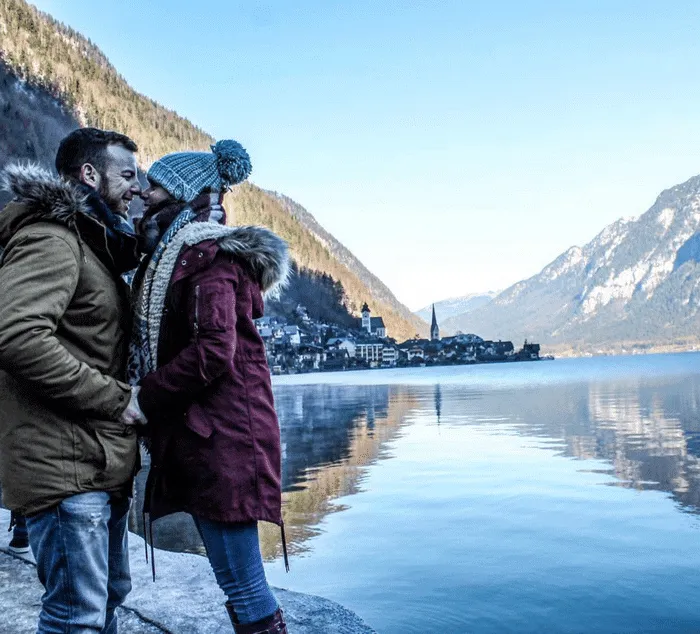 Pareja abrazándose frente a un lago rodeado de montañas y un pueblo pintoresco.