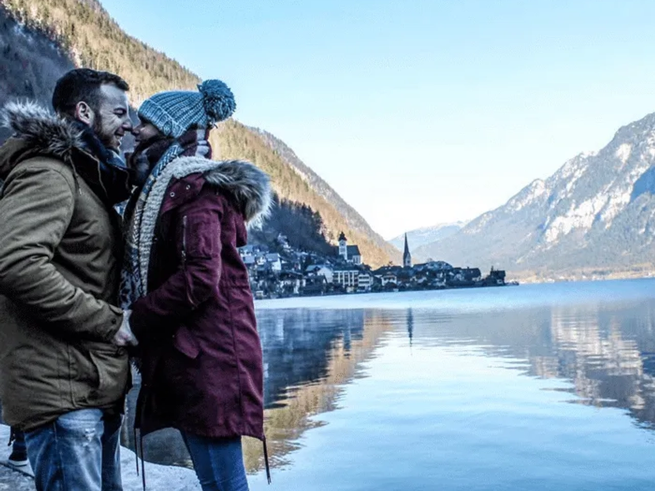 Pareja abrazándose frente a un lago rodeado de montañas y un pueblo pintoresco.
