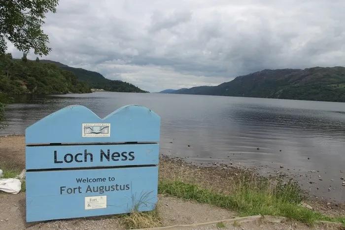 Cartel de bienvenida al Lago Ness, con paisaje montañoso y cielo nublado.
