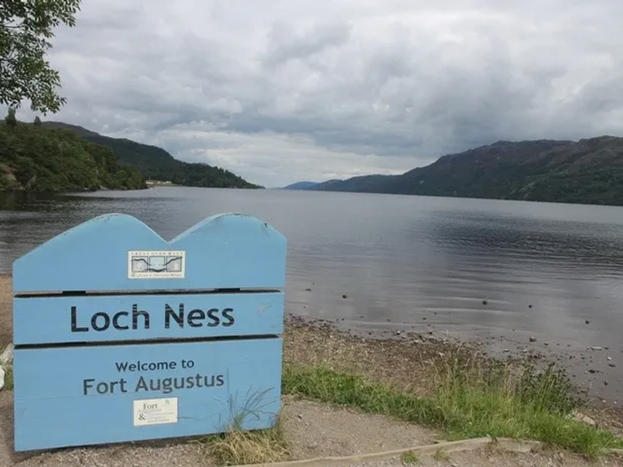 Cartel de bienvenida al Lago Ness, con paisaje montañoso y cielo nublado.