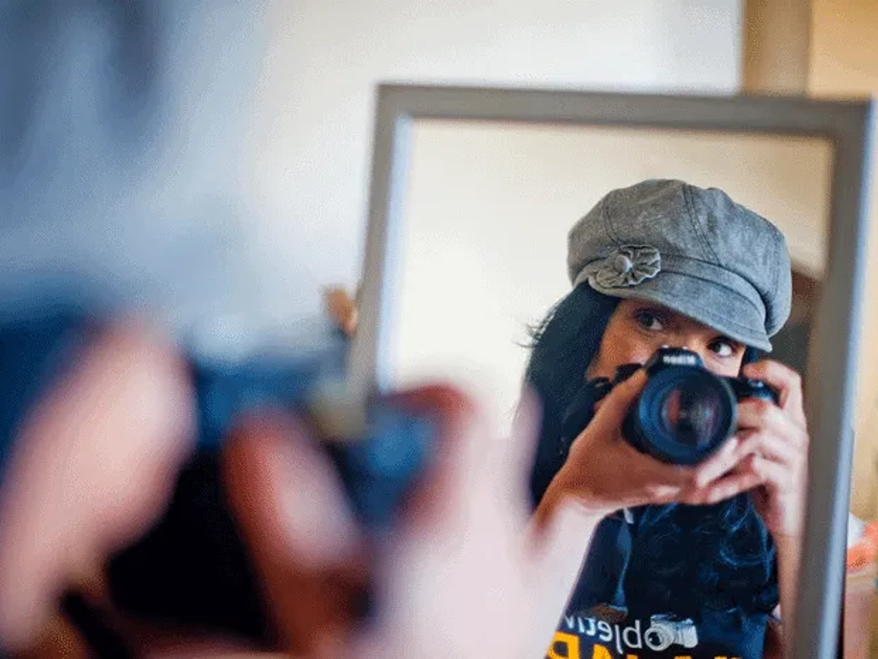 Mujer con gorra se refleja en un espejo mientras sostiene una cámara.