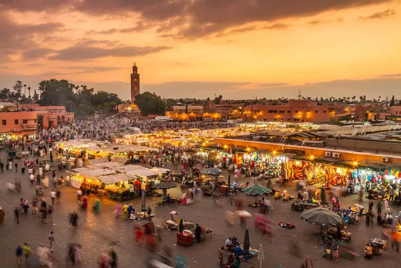 Piazza Jemaa el-Fna a Marrakech al tramonto, con bancarelle illuminate, folla in movimento e la Koutoubia sullo sfondo.