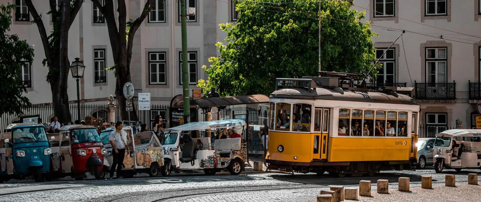 Elétrico amarelo passando por rua movimentada, com tuk-tuks e pessoas ao redor.