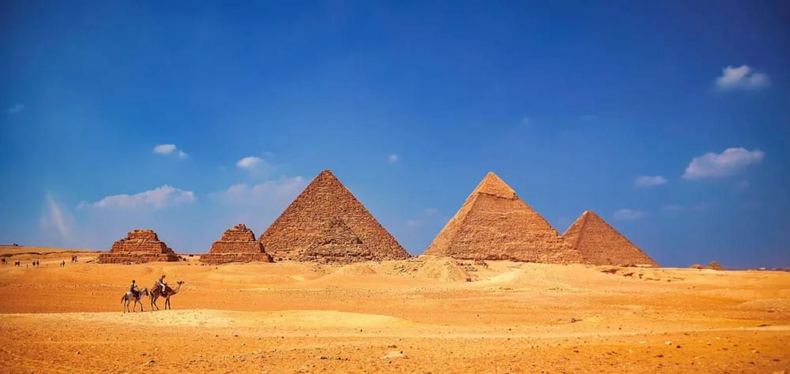 Vista panoramica delle piramidi di Giza nel deserto egiziano, con alcune persone su cammelli in lontananza sotto un cielo azzurro.