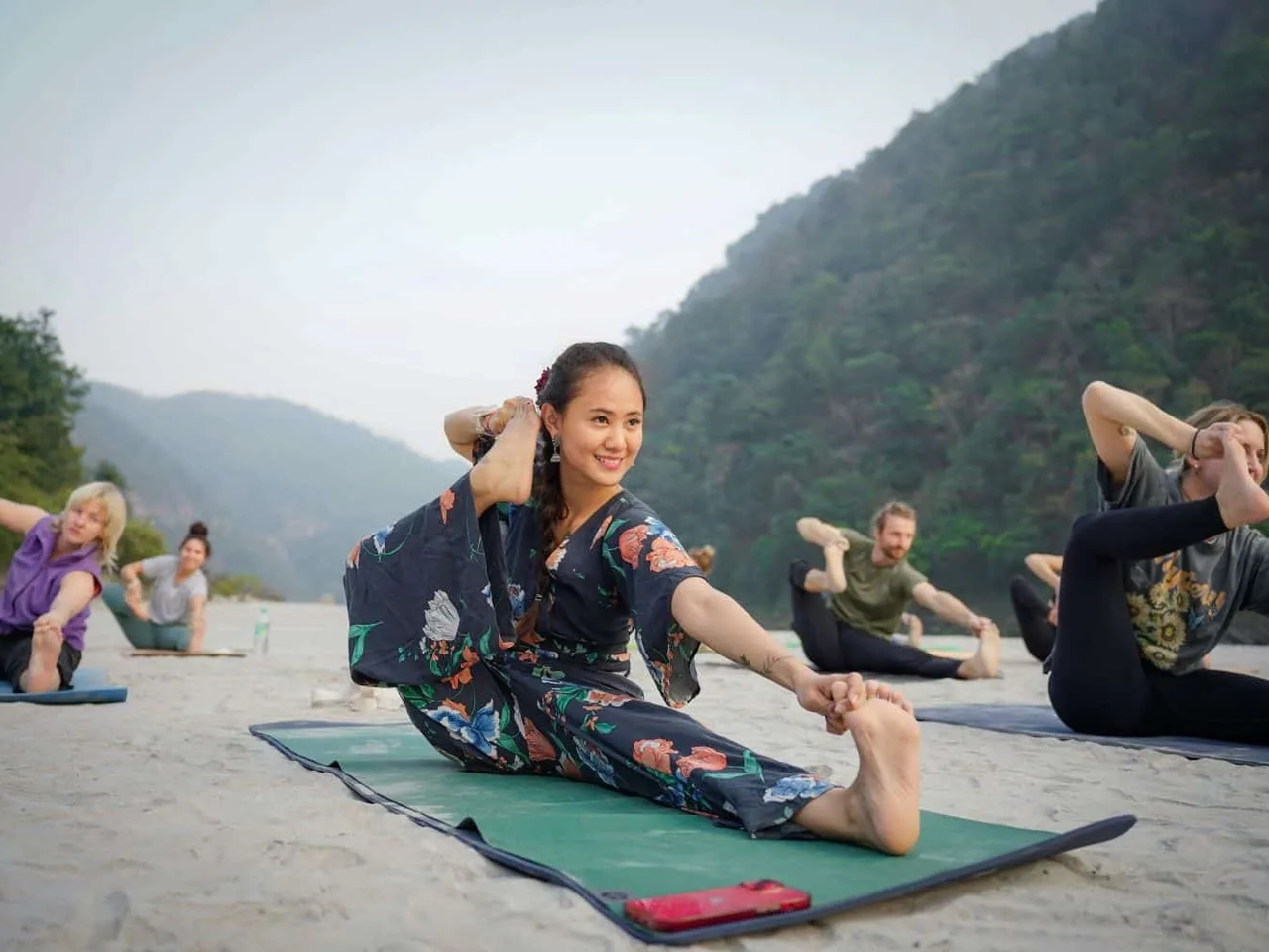 Grupo de pessoas praticando yoga em um ambiente natural, com montanhas ao fundo.