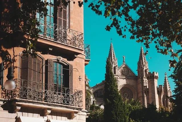 Edifício histórico com janelas de ferro e catedral ao fundo em dia ensolarado.