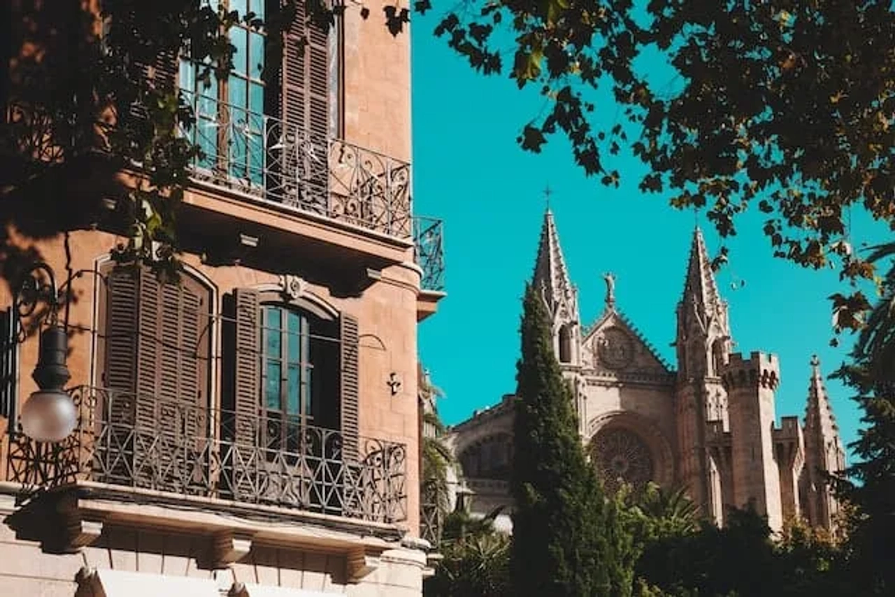 Edifício histórico com janelas de ferro e catedral ao fundo em dia ensolarado.