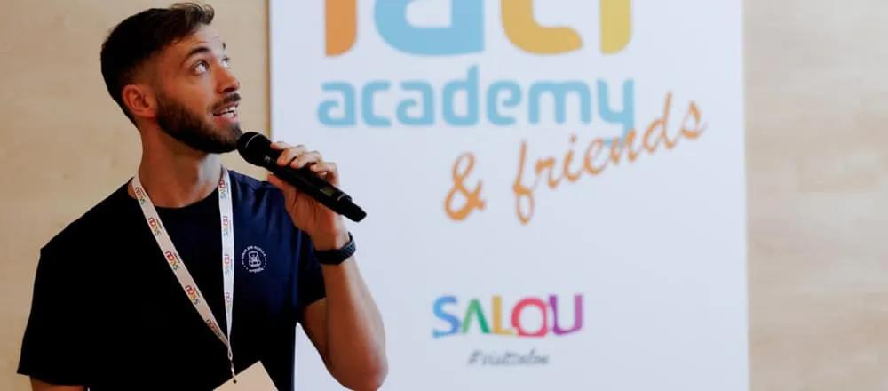 Hombre hablando con micrófono frente a un cartel de IATI Academy & Friends.