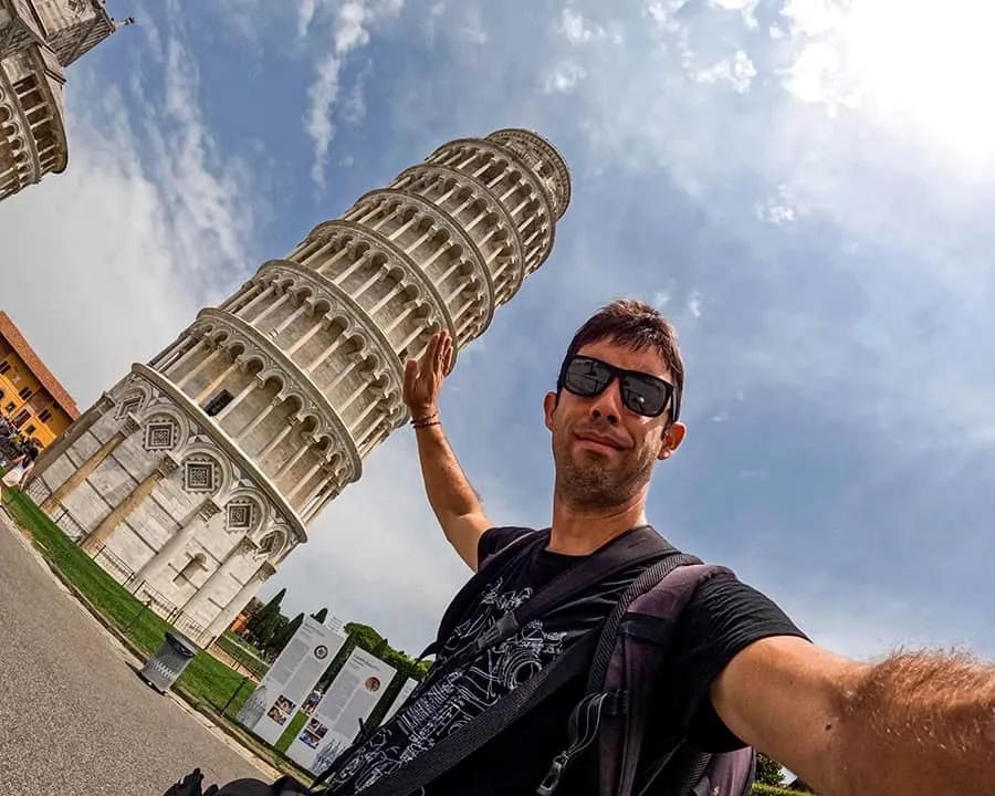Hombre sosteniendo la Torre de Pisa con una mano, fondo de cielo nublado.