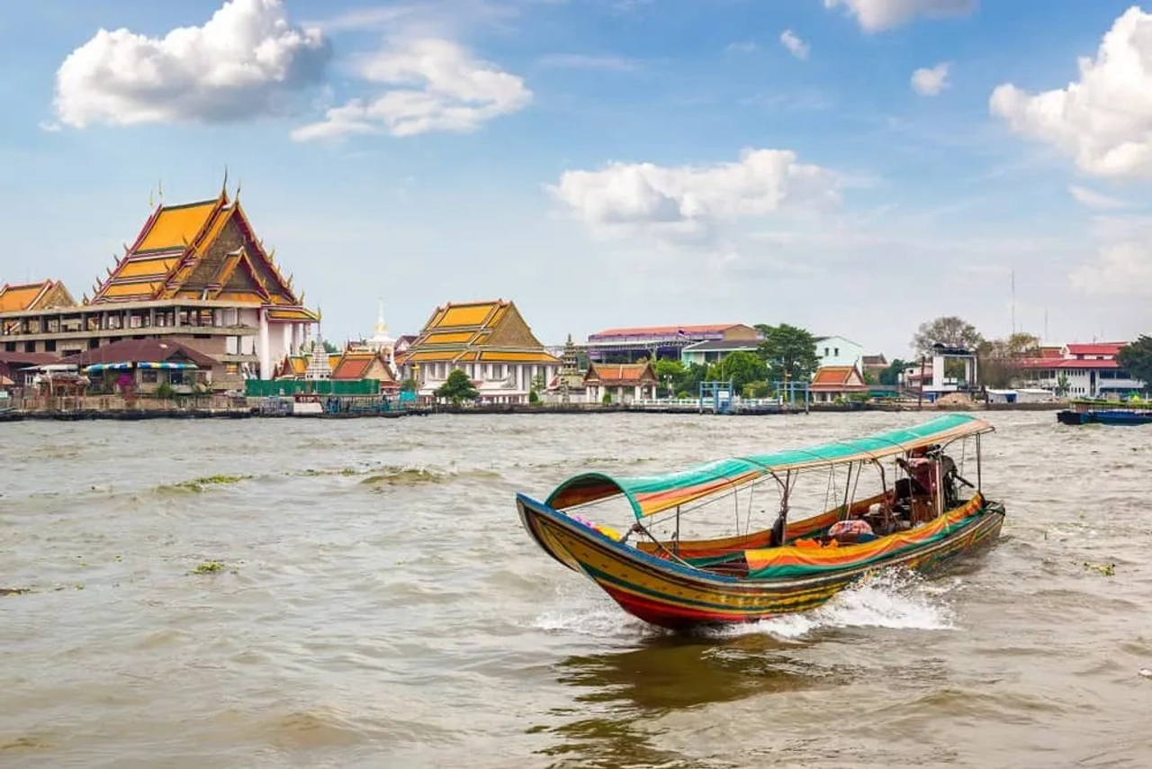 Barca tradizionale sul fiume Chao Phraya a Bangkok, con templi thailandesi sulla riva e cielo azzurro sullo sfondo.