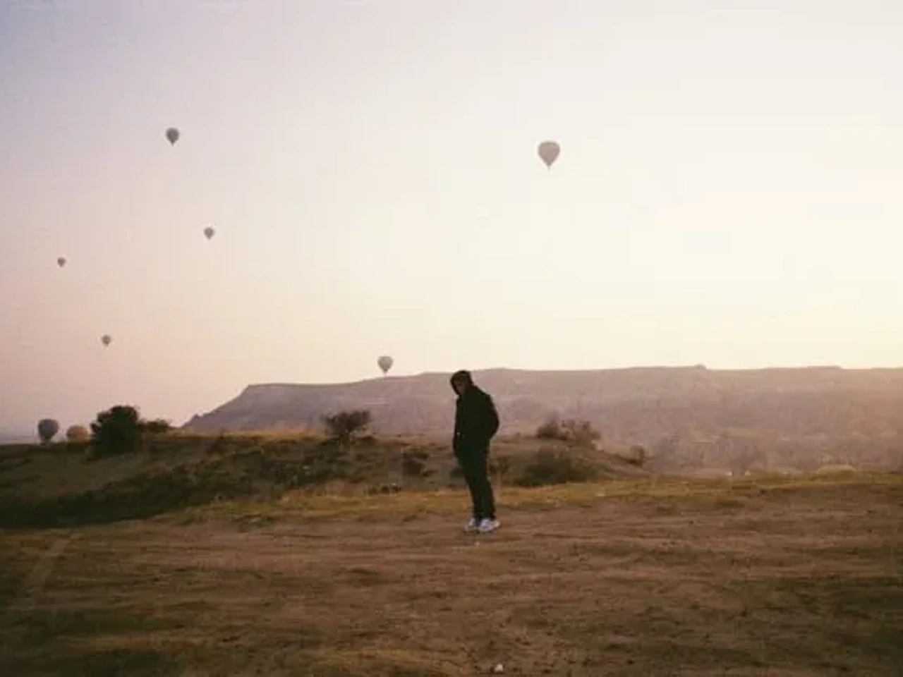 homem com um casaco e as mãos nos bolsos junto a baloes de ar quente na capadocia na turquia