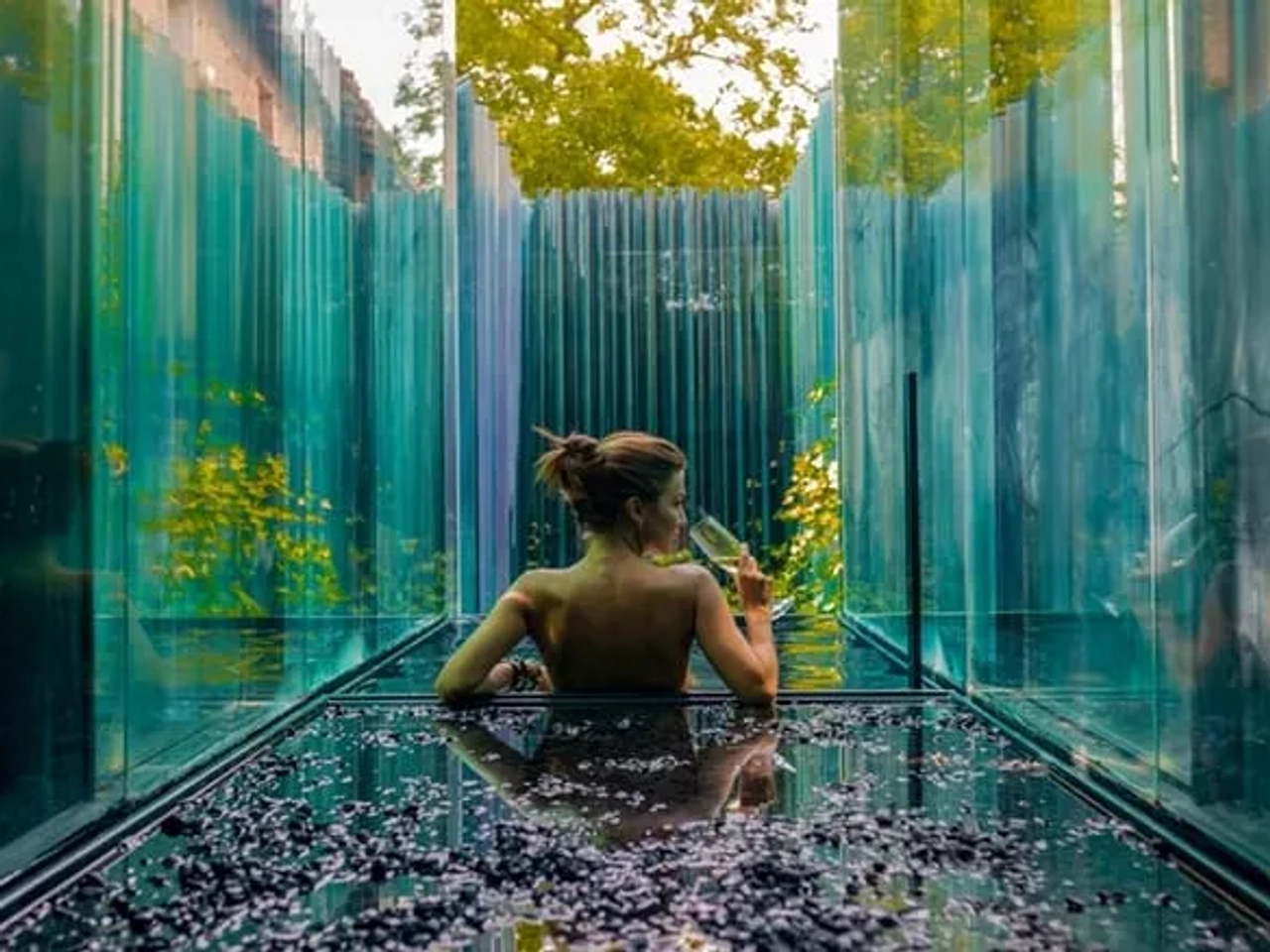 Mujer relajándose en un baño de vidrio rodeado de naturaleza.