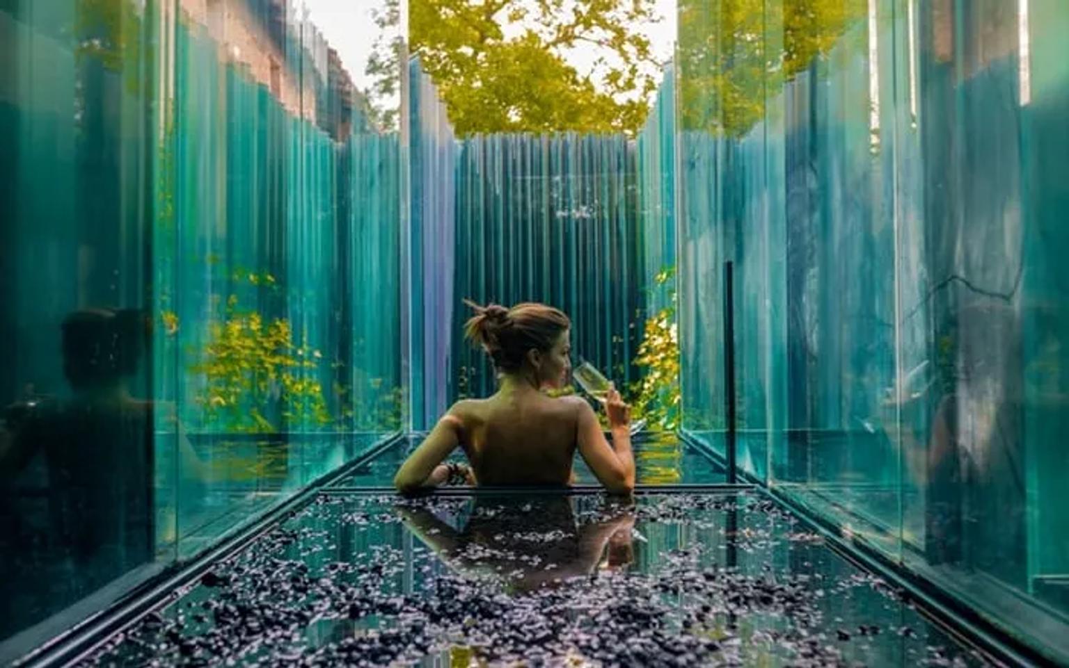 Mujer relajándose en un baño de vidrio rodeado de naturaleza.