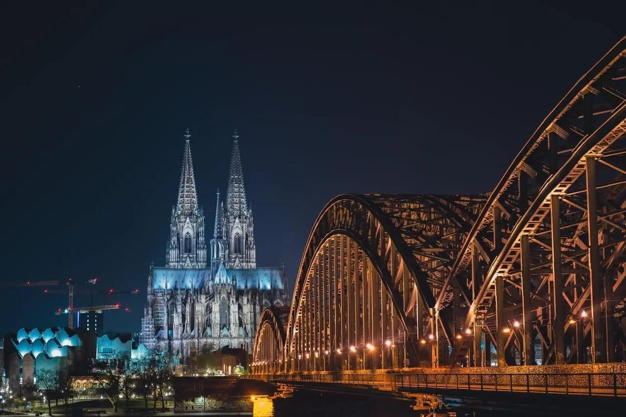 Catedral iluminada à noite com ponte metálica em primeiro plano.