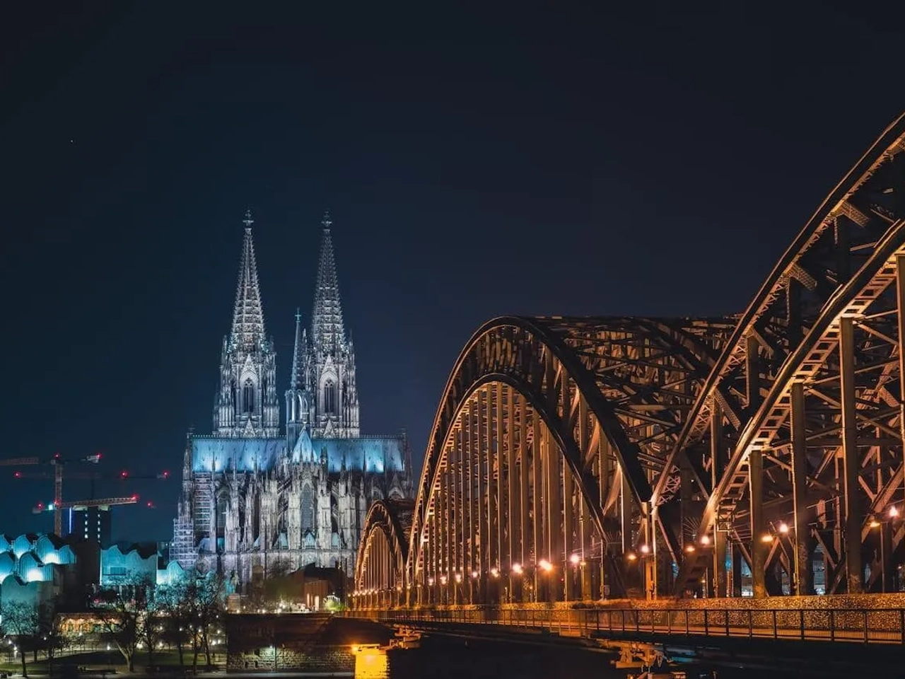 Catedral iluminada à noite com ponte metálica em primeiro plano.