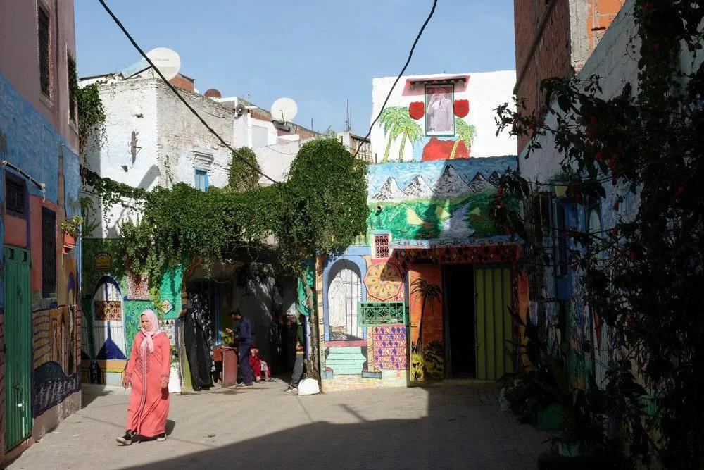Calle colorida en Marruecos con murales y vegetación, una mujer camina.