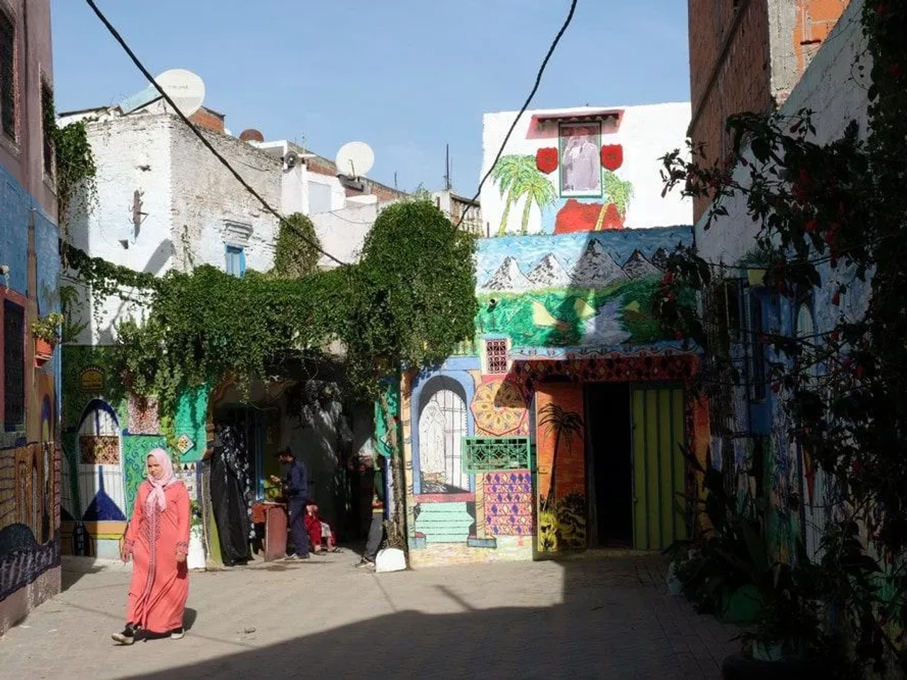 Calle colorida en Marruecos con murales y vegetación, una mujer camina.