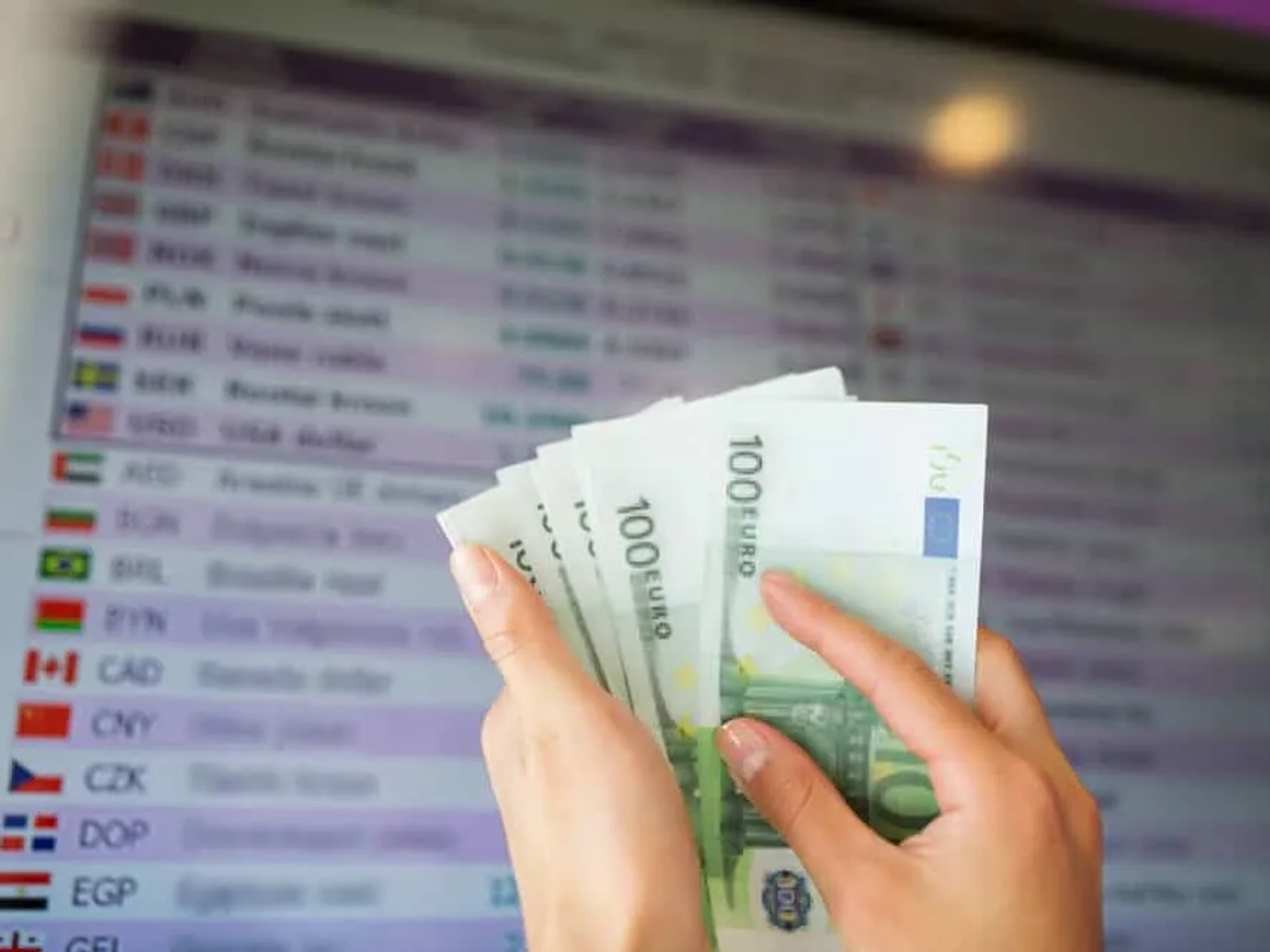 pessoa com notas de 100 euros e placard de taxas de cambio