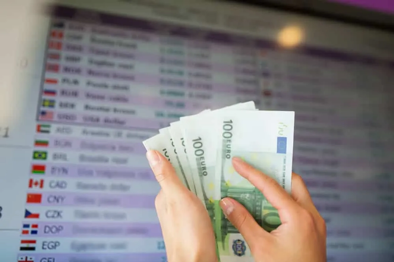 pessoa com notas de 100 euros e placard de taxas de cambio