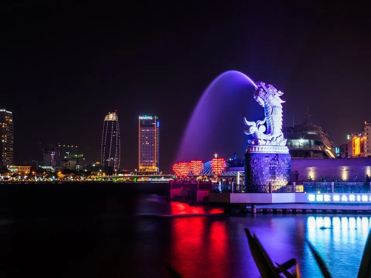Fonte iluminada em forma de dragão à noite, com skyline da cidade ao fundo.