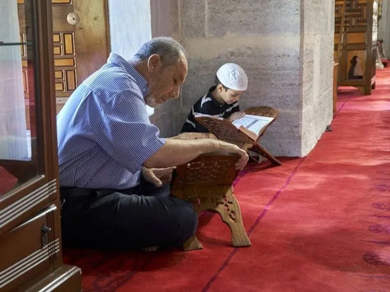 Homem e criança lendo em uma mesquita, com tapete vermelho ao fundo.