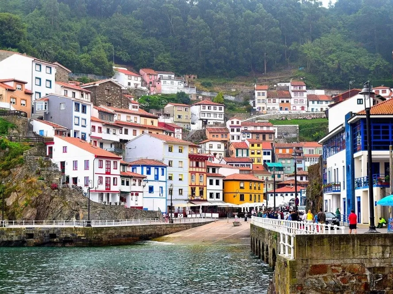 Vista de casas coloridas à beira-mar nas Astúrias, com pessoas caminhando.